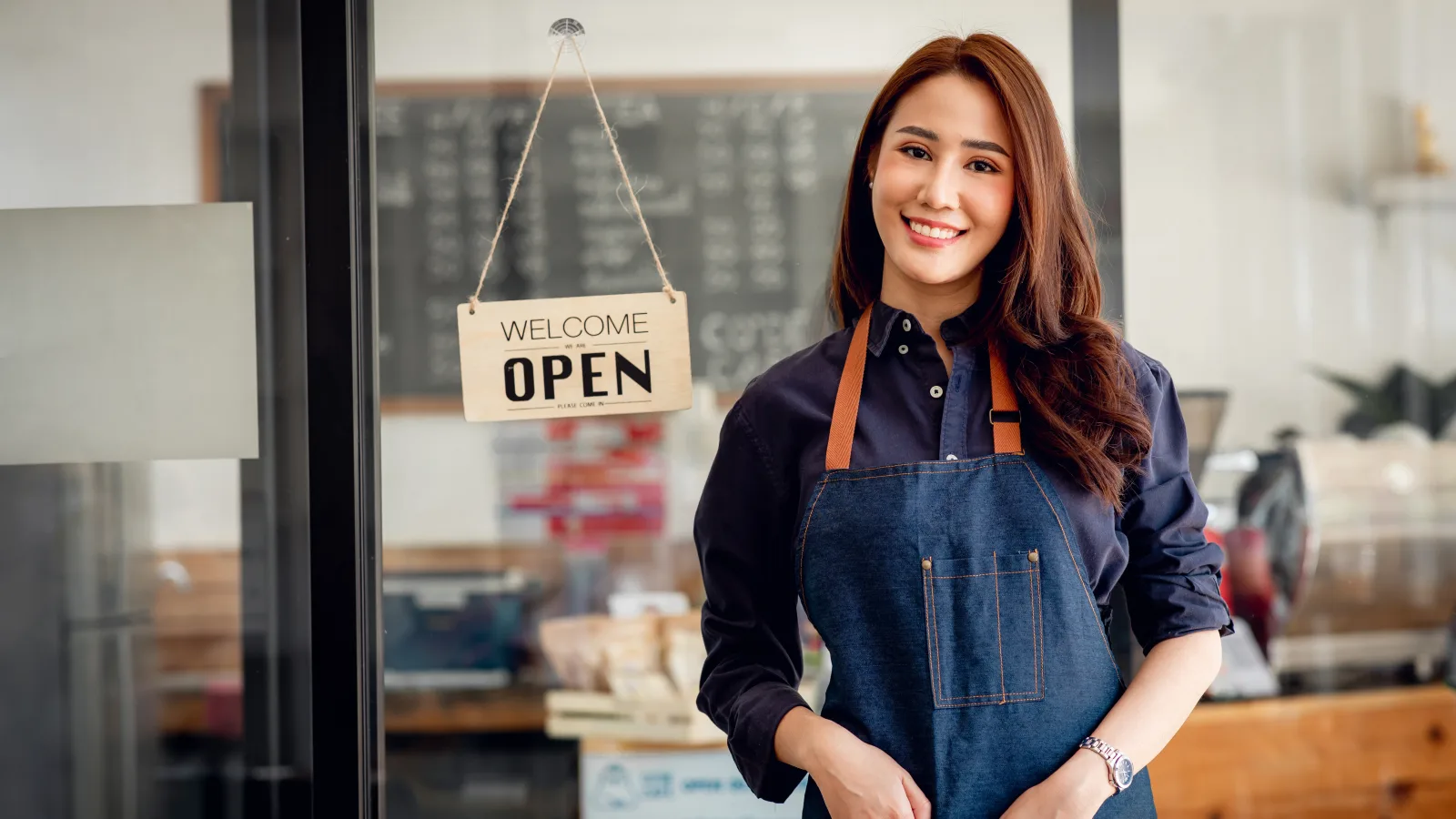 Ladeninhaberin mit Schürze vor ihrem Geschäft, im Hintergrund ein Schild mit der Aufschrift „Open